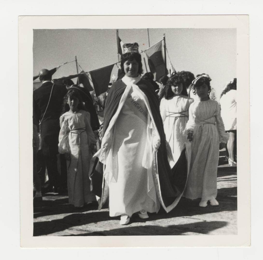 Young woman dressed in a long gown, cape and crown. Three younger women stand behind her holding the end of her cape off the ground