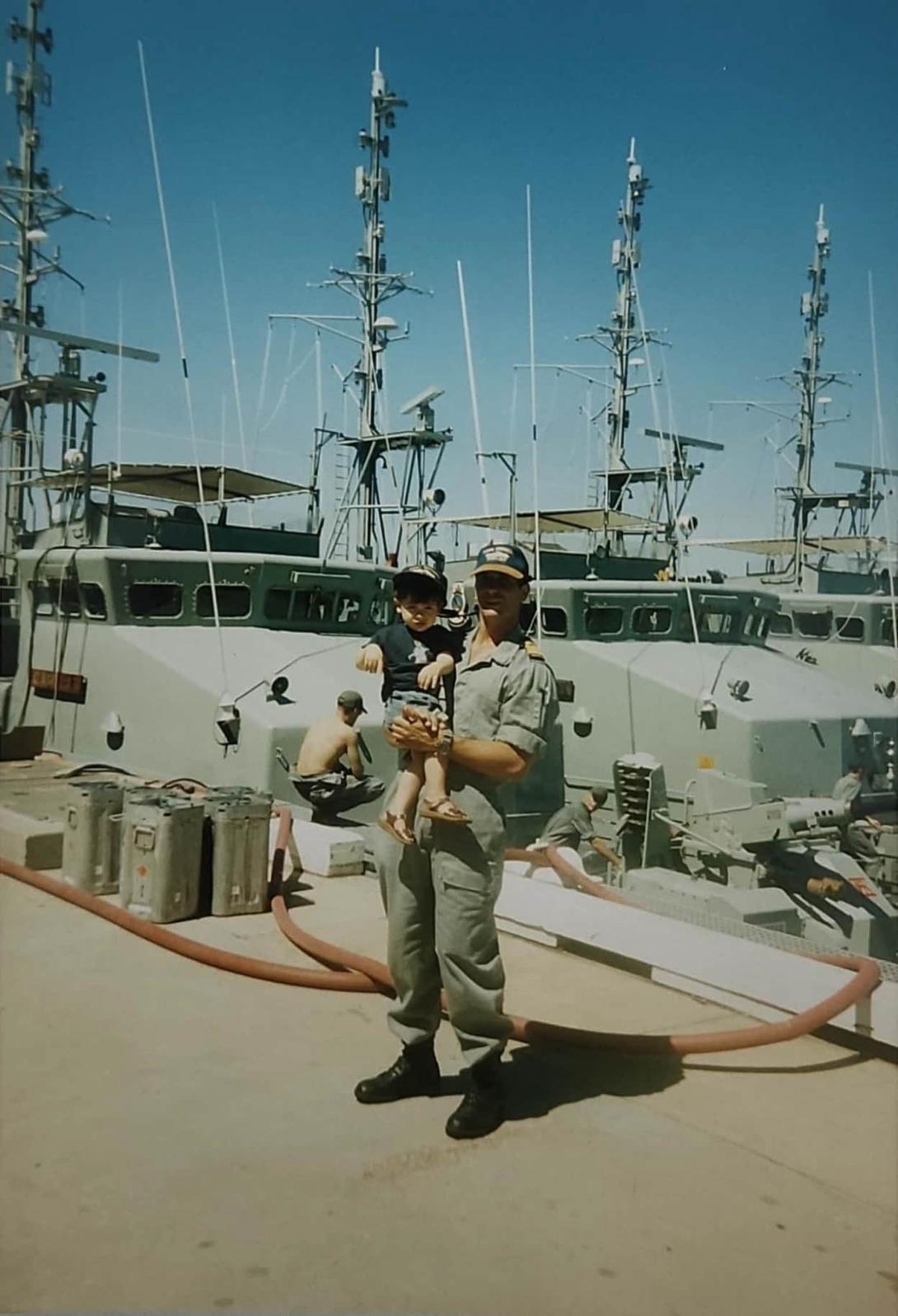 Paul in a plain uniform holding up a small child in front of a row of armoured naval boats.