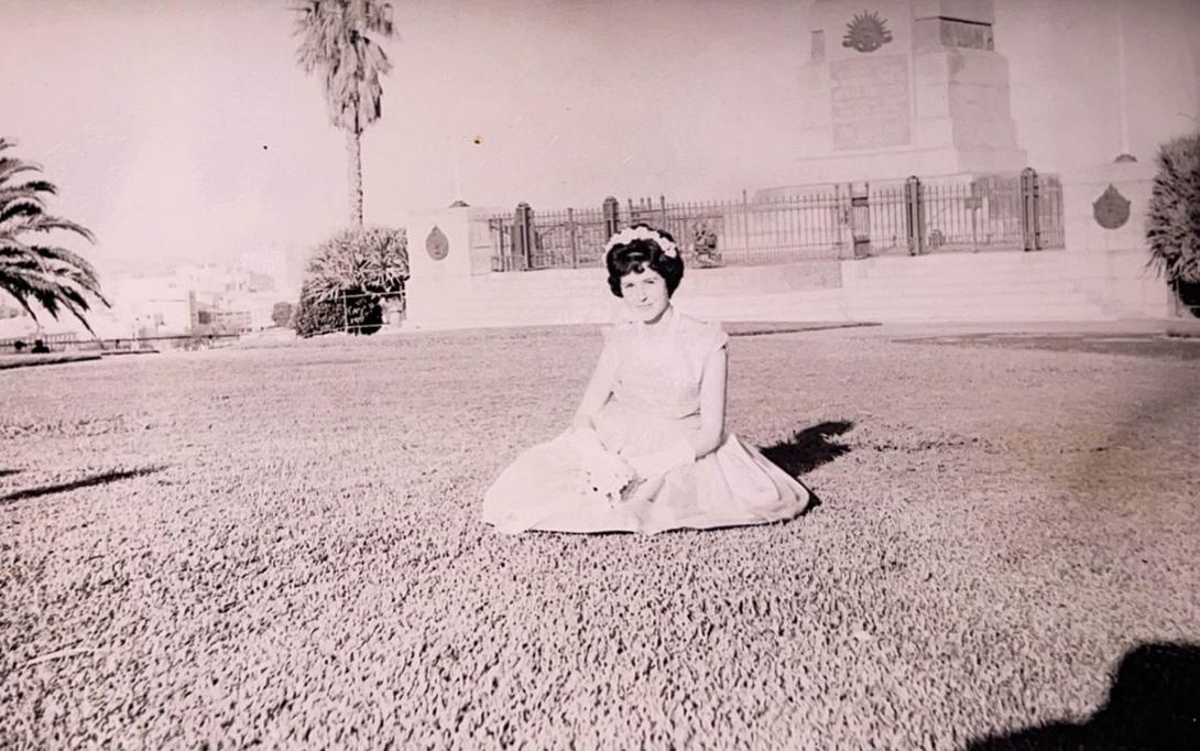 Carmela in a white dress seated in a large grassy park.