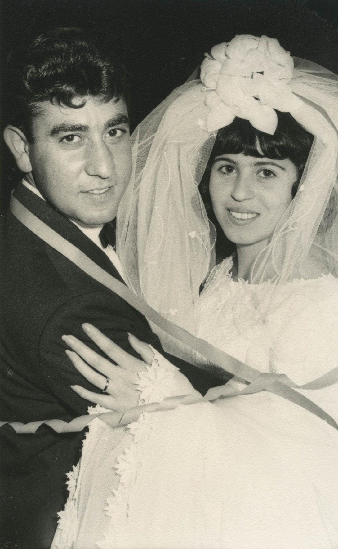 Carmela and Santo embrace, he is in a black suit and she is in a wedding dress and veil.