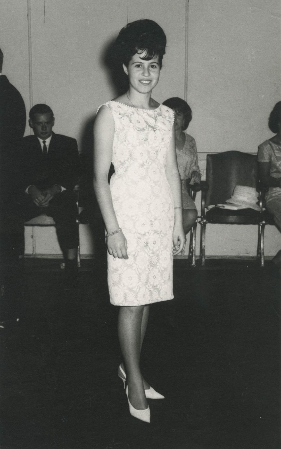 Carmela in a short white sleeveless dress, standing at a party.