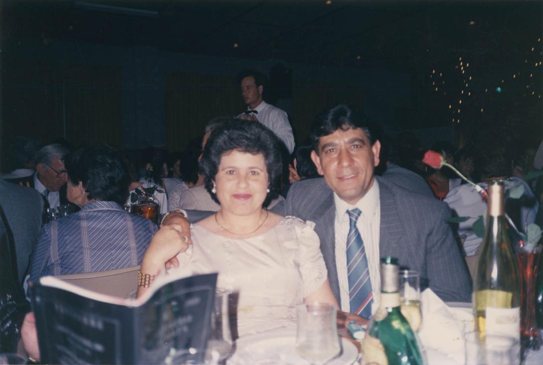 Santo's arm around Carmela's shoulder at a table.
