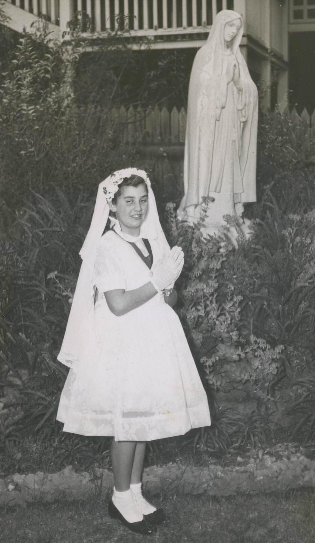 Louisa stands in white dress in veil in front of a statue of the Madonna.