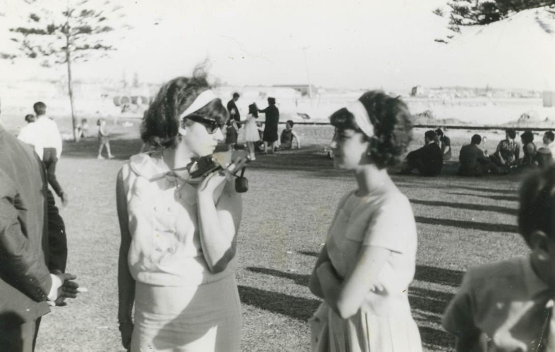 The sisters chat in a grassy field among crowds of people enjoying the sunny day.