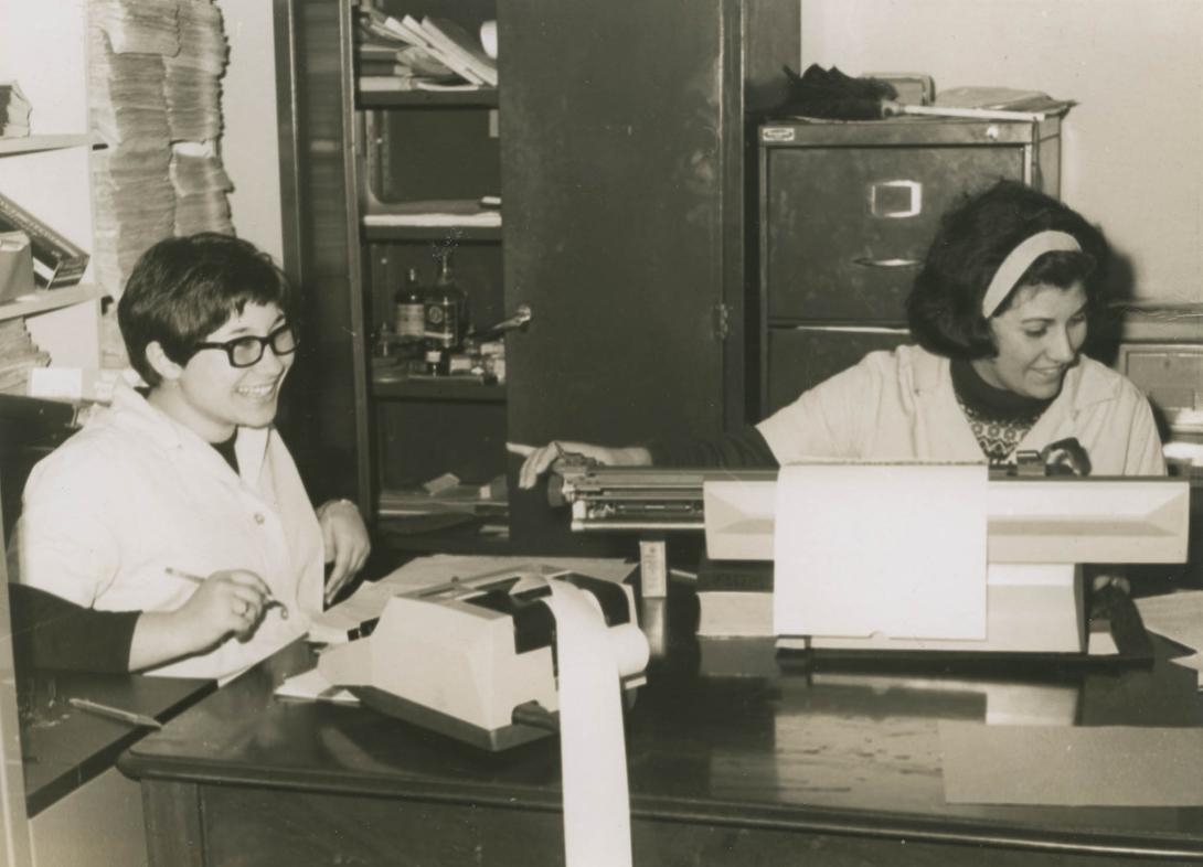 Two young women enjoying their work at a giant ticker-tape calculator and other old office equipment.