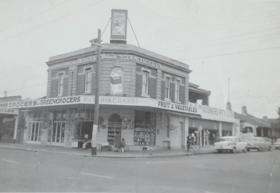 The large two-storey corner store advertises fruit & vegetables, Carbonell cigars and Capstan cigarettes.