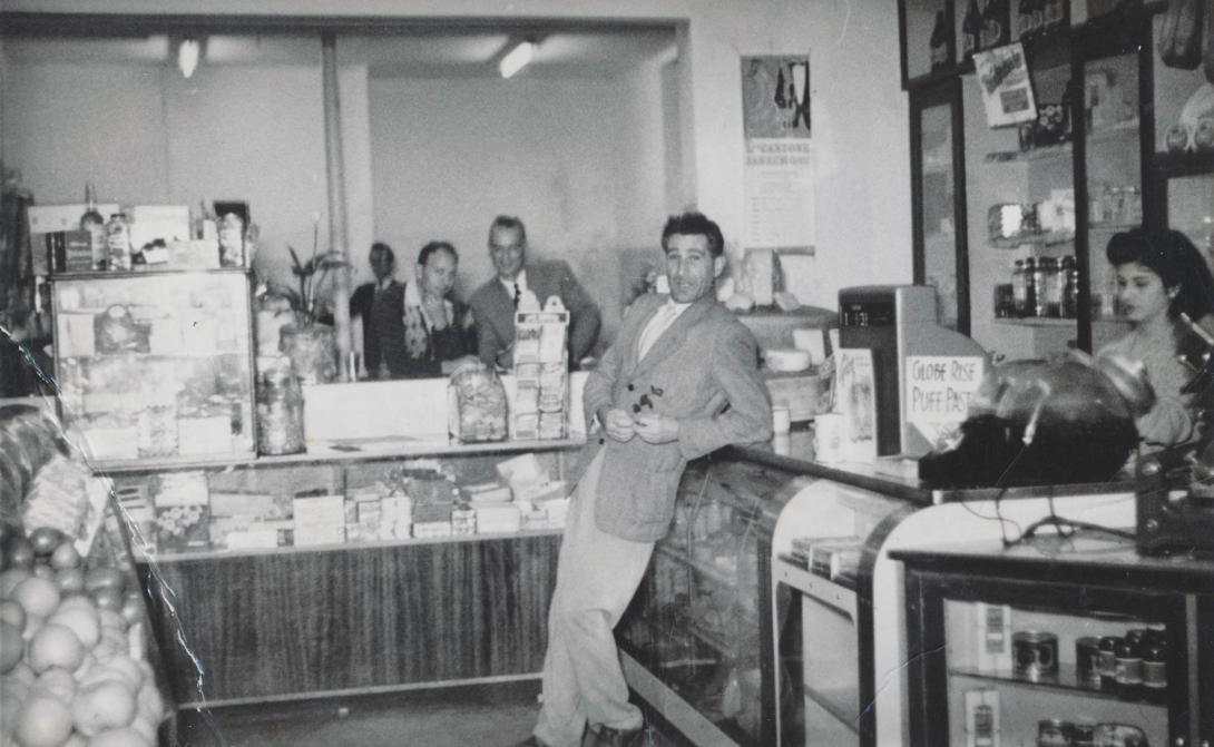 A well-dressed man leans back jovially against the counter where Betty is serving.