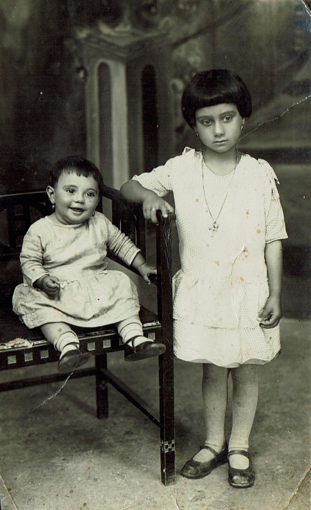 A baby on a chair looking very happy, alongside an older child standing with a very serious expression.
