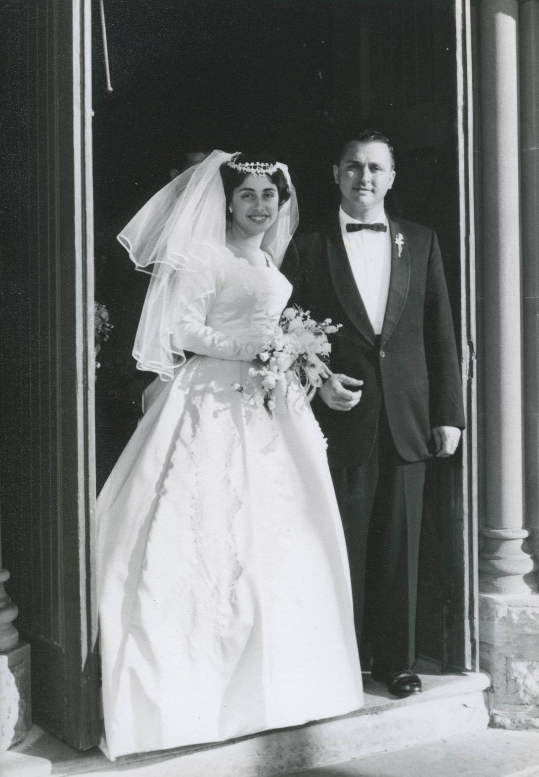 A beaming Mary in a white wedding gown and veil, her hand resting on the arm of her proud husband.