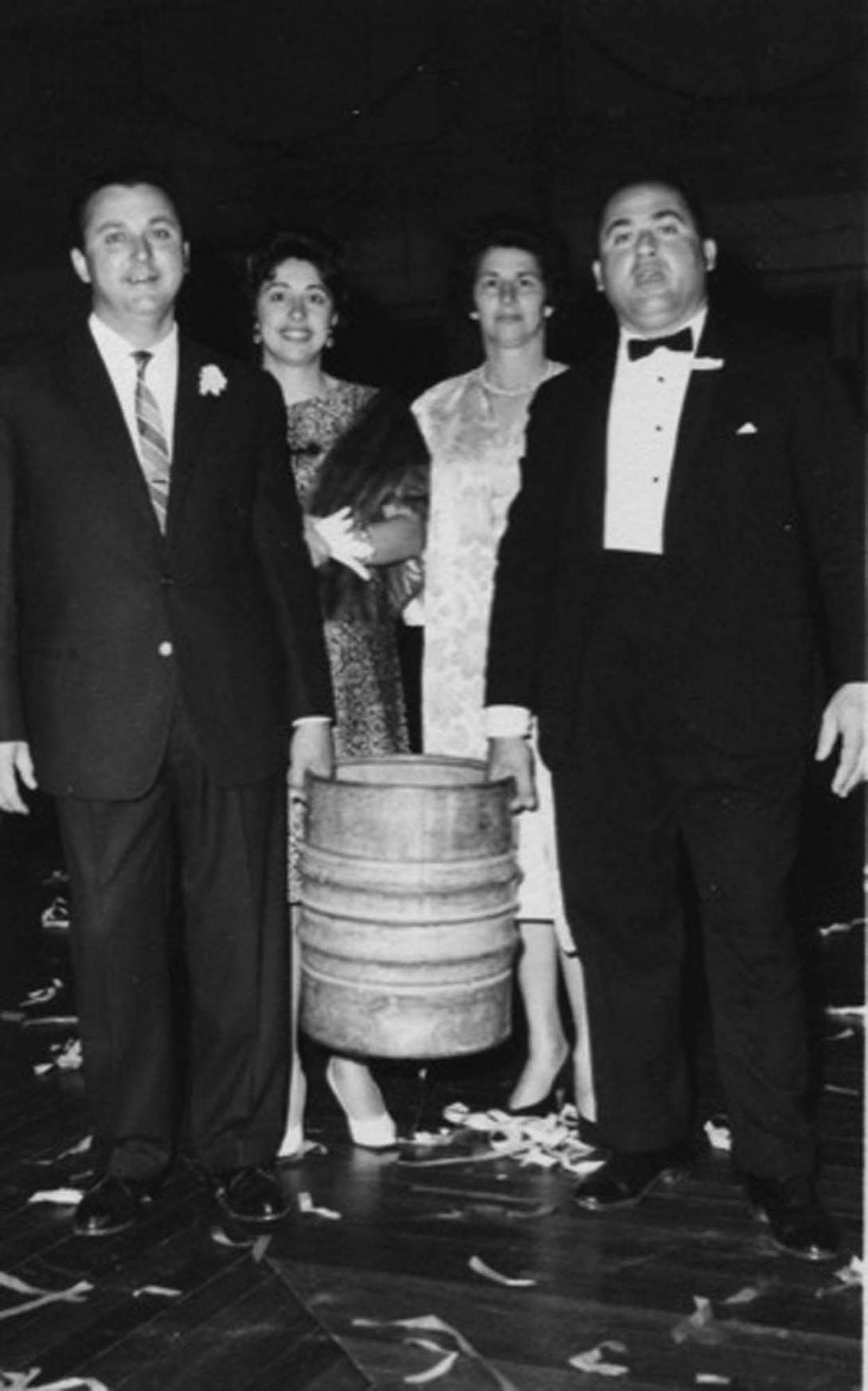 The two men in black suits and bow ties lift a heavy metal beer barrel between them. The two women stand behind.