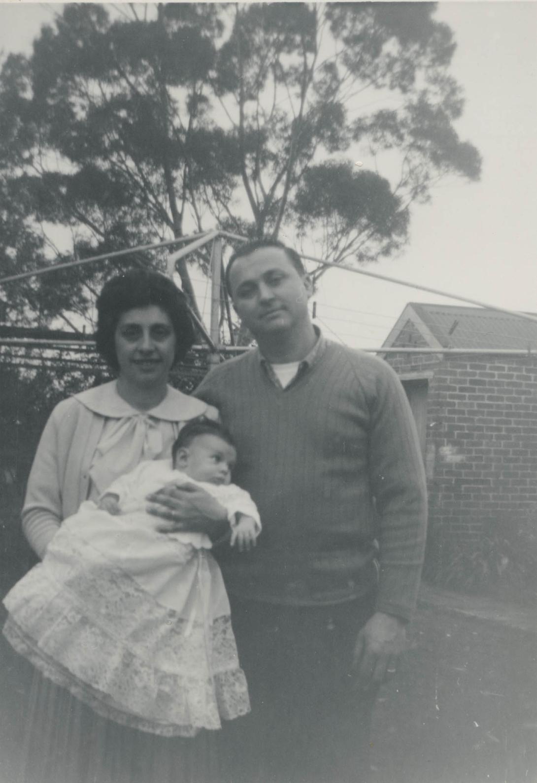 In a suburban backyard, the couple with a baby in a white dress stand in front of a Hills Hoist.