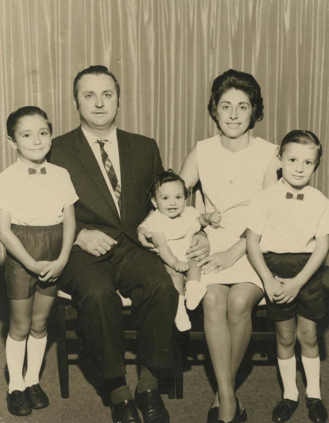 The two older boys wear white shirts, shorts and bow ties with tall white socks flank their parents and the baby.