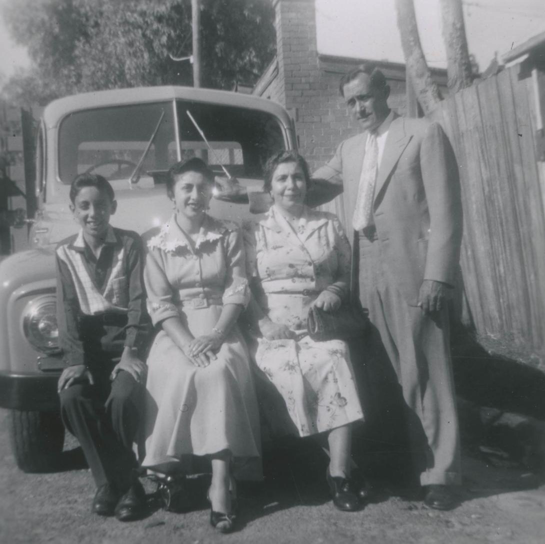 Two women and a boy perched on the bumper of a shiny old model car. A man in a pale suit stands with a hand on the bonnet.