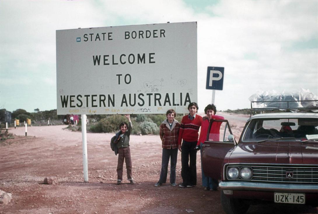 Graffiti marks the lower half of a large white sign reading "Welcome to Western Australia." The family stand beside it and their shiny car.