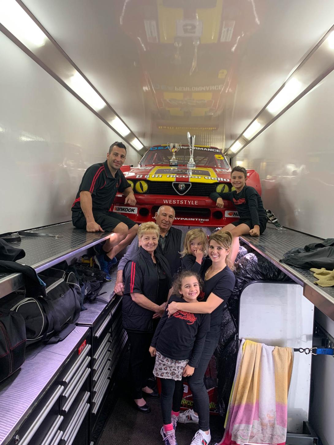 Everyone's crammed in a narrow truck trailer, with the racing car raised up high at the back.