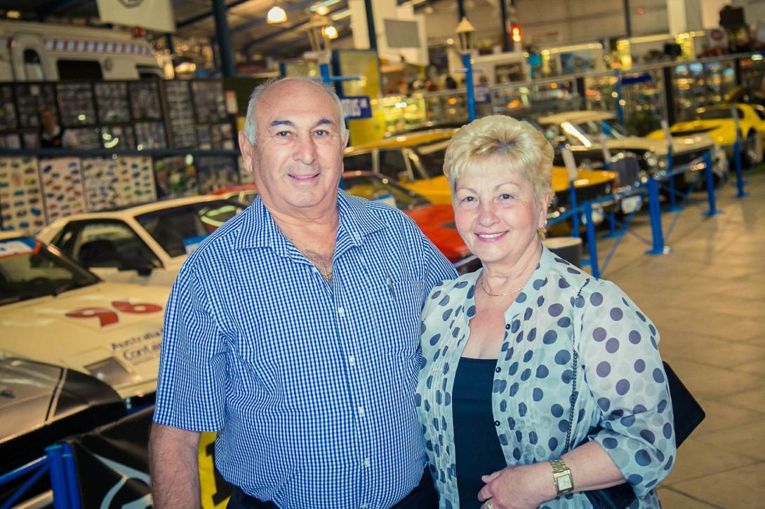 The couple stand together in front of many shiny racing cars.