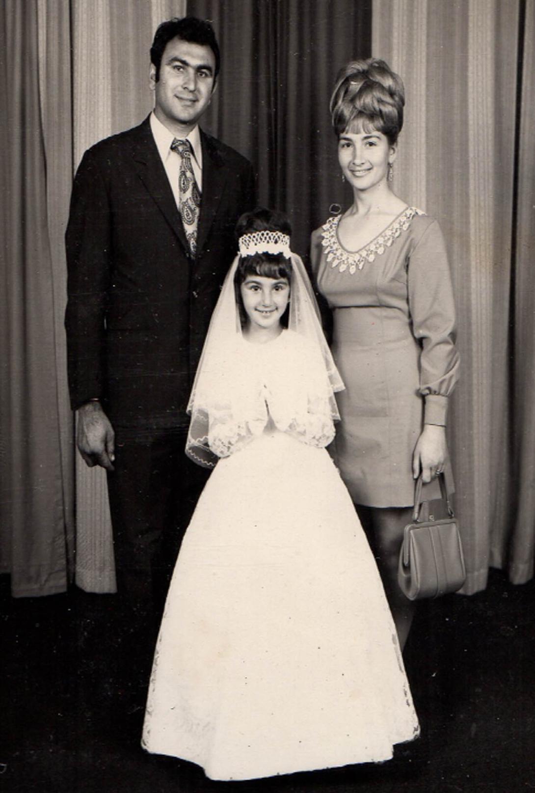 A smiling Rosemary in long white dress and headress, with parents behind.