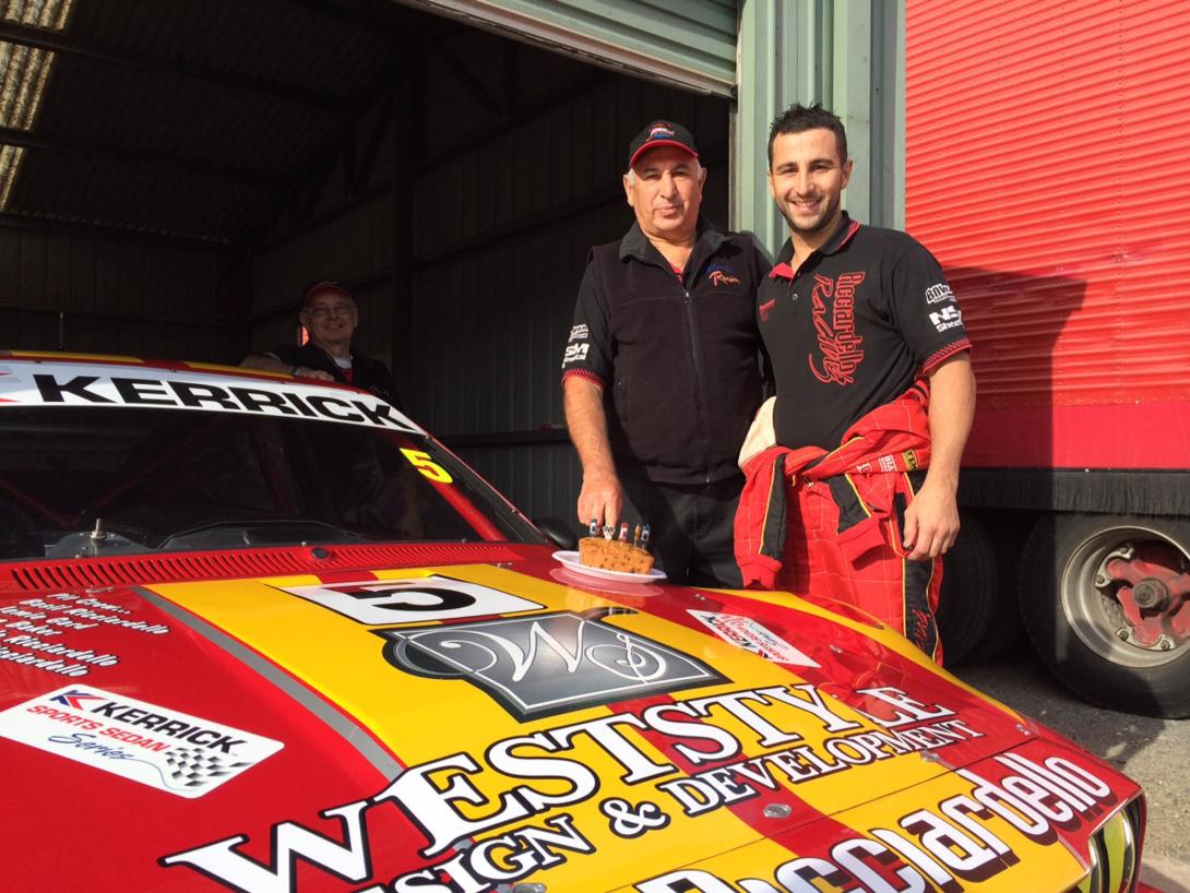 Dad & son stand behind a brightly coloured racecar covered in sponsorship logos.