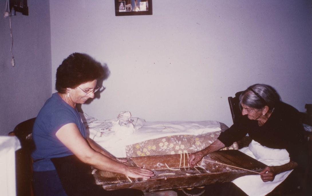The two women bend over a long wooden tray working the pasta.