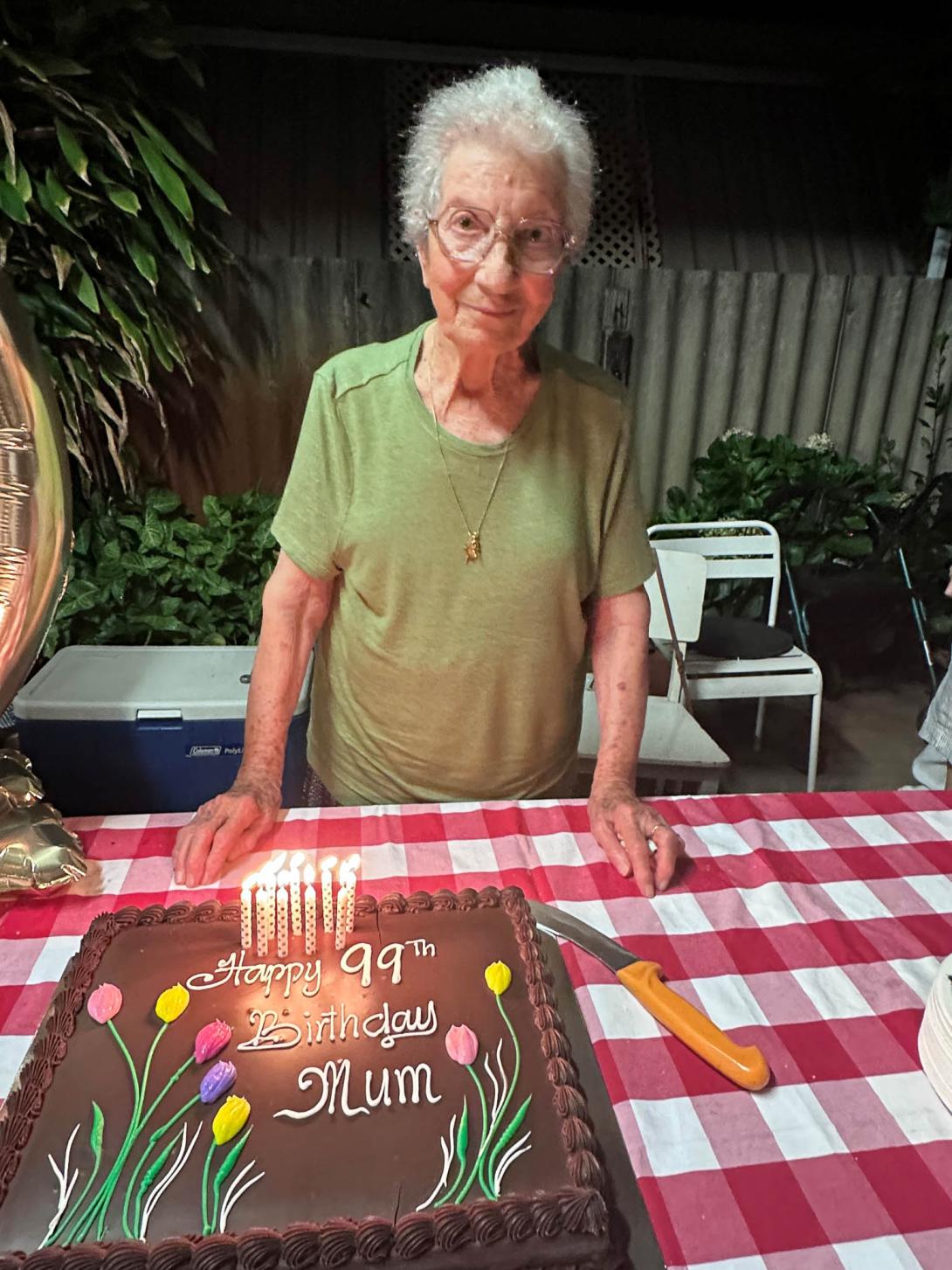 Paola behind a big chocolate cake reading "Happy 99th Birthday Mum." There are some candles in the cake, but there's not 99 of them.