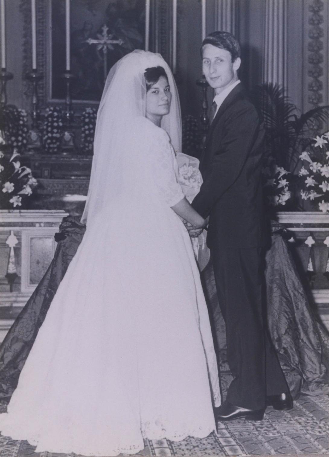 Franca in a long white dress and veil holds hands with Fabrizio in front of fine church decorations.