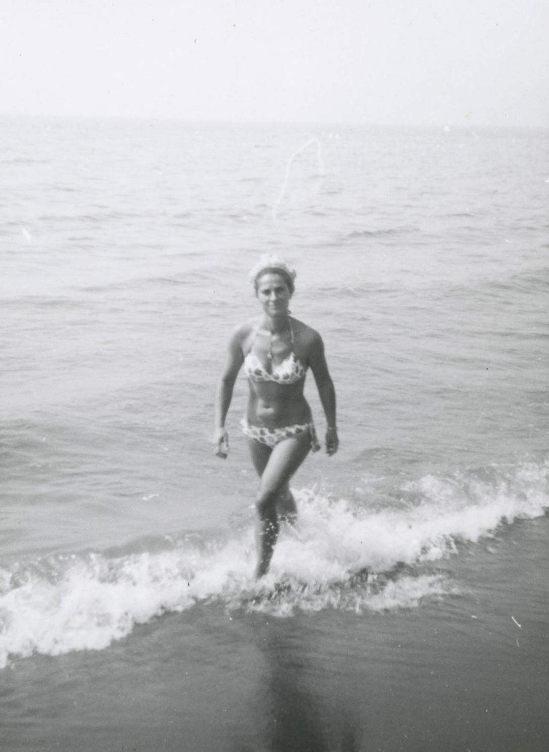 Franca wears a patterned bikini, ankles in the surf.
