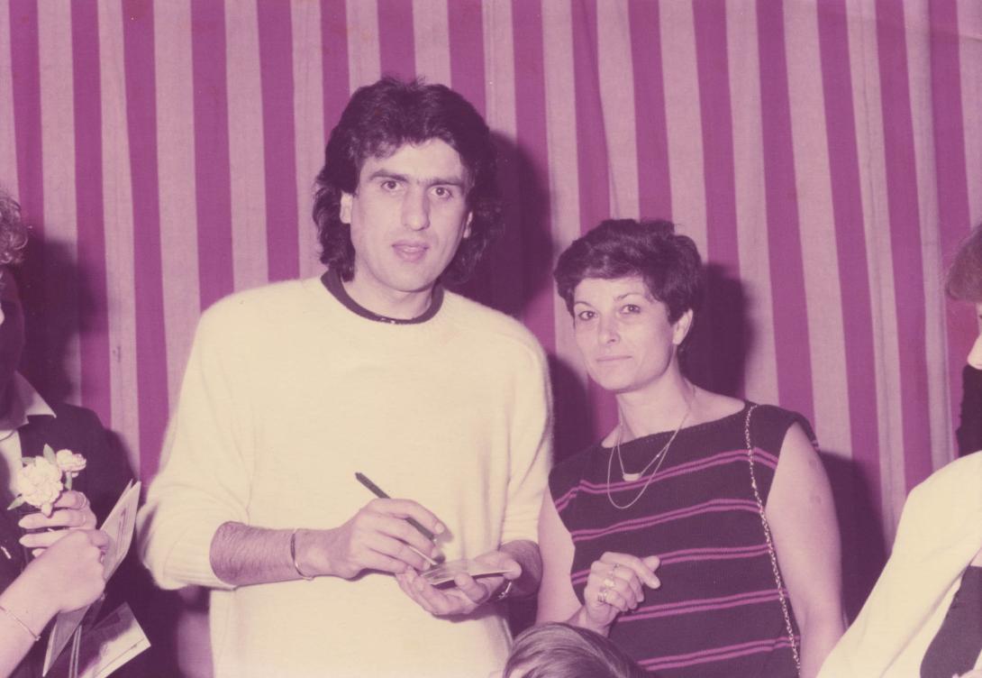 Franca stands with Tony who's in a bright yellow shirt in front of a backdrop striped pink and purple.