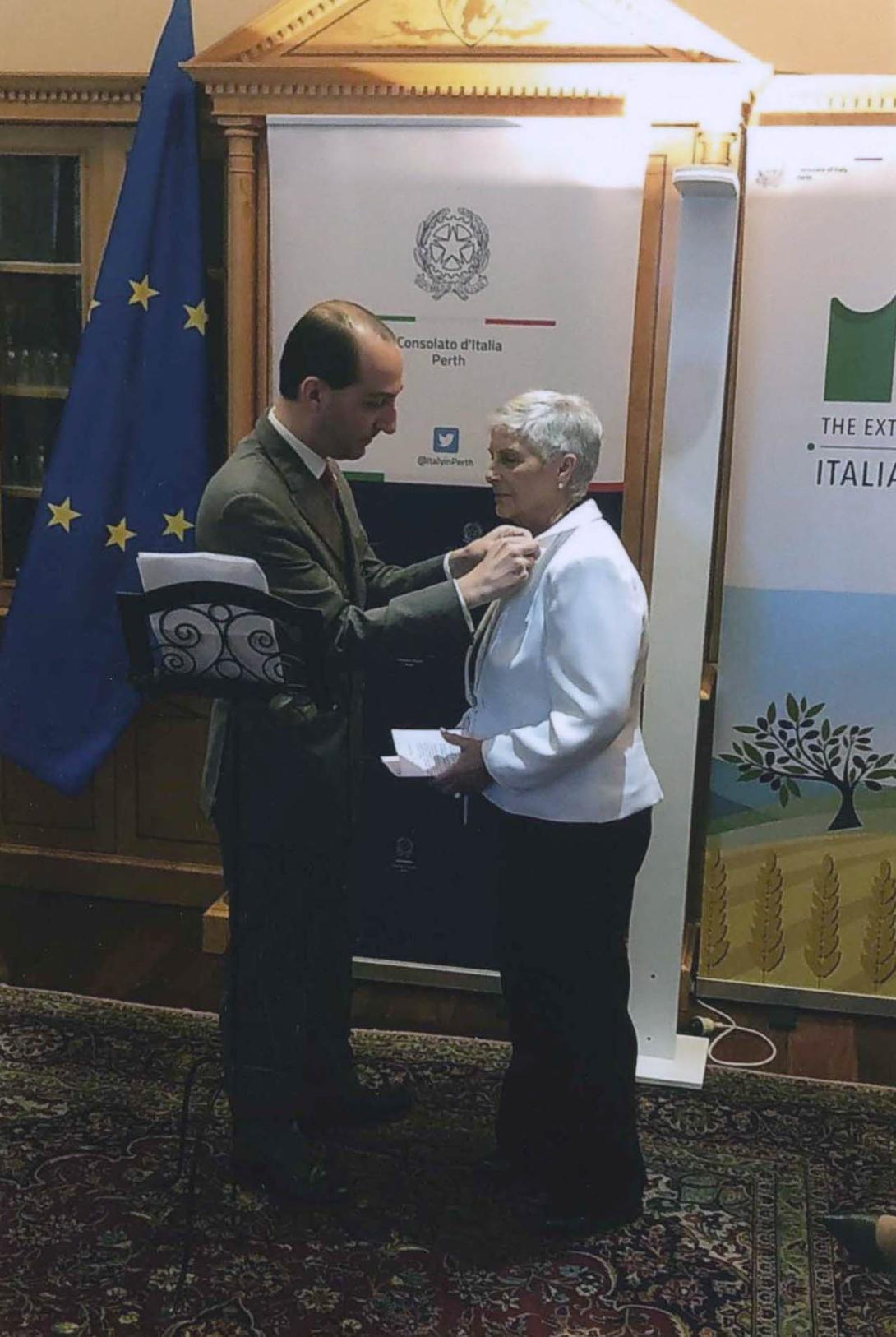 In front of a banner reading "Consolato d'Italia Perth," the Consul pins the star to Franca's lapel.