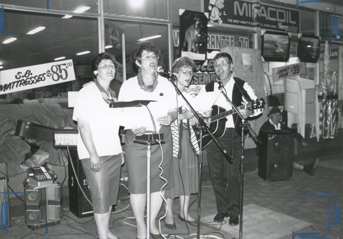 A quartet singing, one playing a guitar in front of a store advertising mattresses for $85.