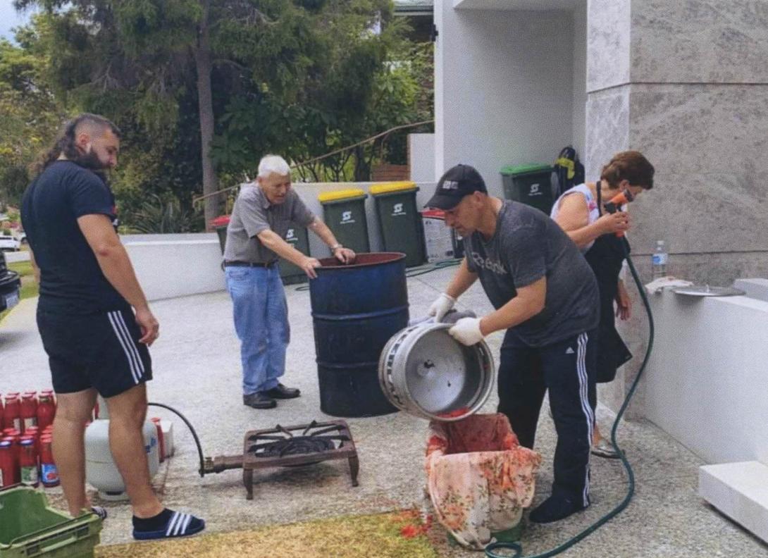 The family outdoors with a large barrel, keg and gas burner on the ground. A lot of sauce is being made.