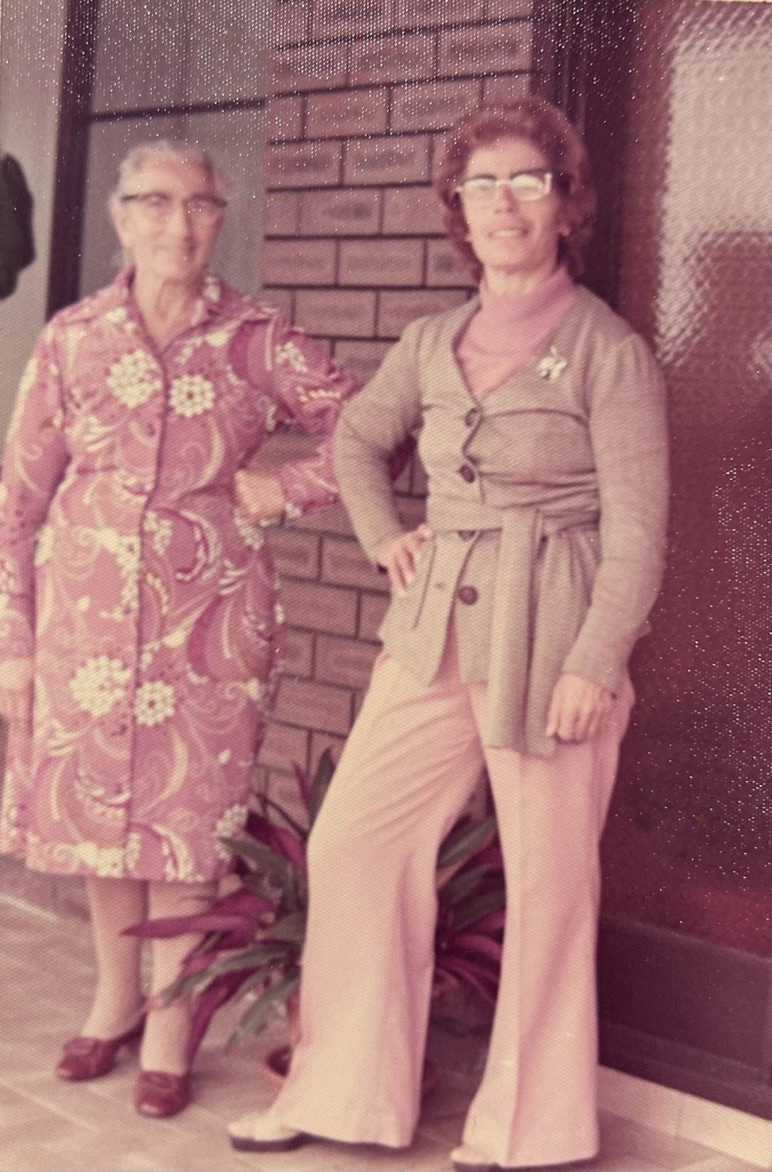 Carolina in stylish 70s clothing stands alongside her mother in front of red bricks and a pot plant.
