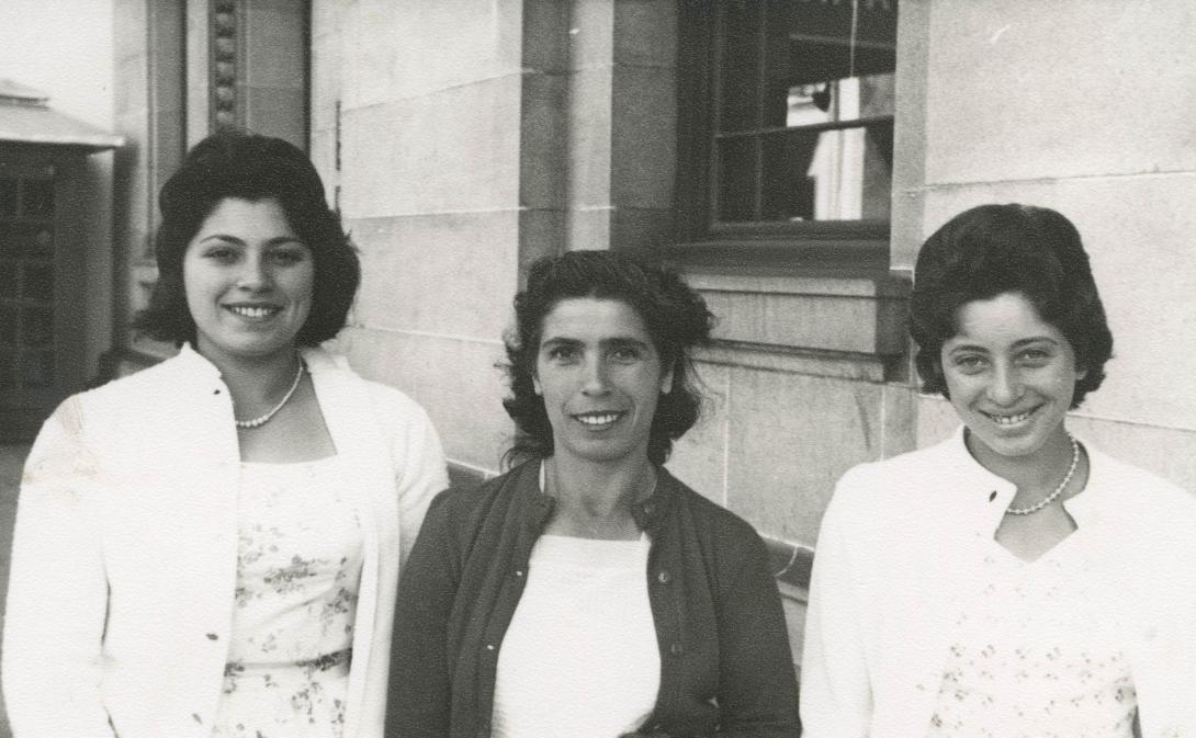 The daughters in white dresses, jackets and shoes stand behind their mother in a darker jacket and skirt.