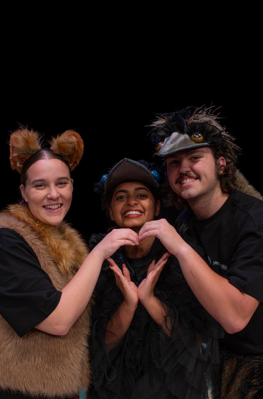 Three performers from Yirra Yaakin' 2026 production of Djinda Kaatijin, shown together in costume (Emu, Crow and Dingo) all smiling and making a loveheart shape with their six hands together.