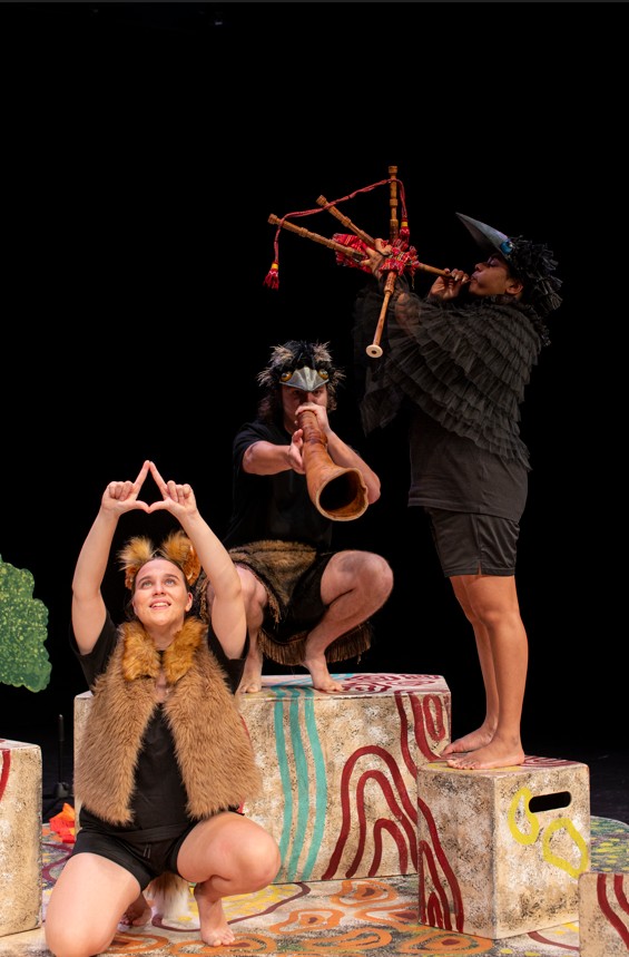 Yirra Yaakin Theatre Company present Djinda Kaatijin. This production image shows a trio of young Aboriginal theatre performers, one seated with her hands in a star formation above her head; one crouched nearby on a stage block playing didgeridoo, and the third performer standing on a stage block playing the bagpipes.