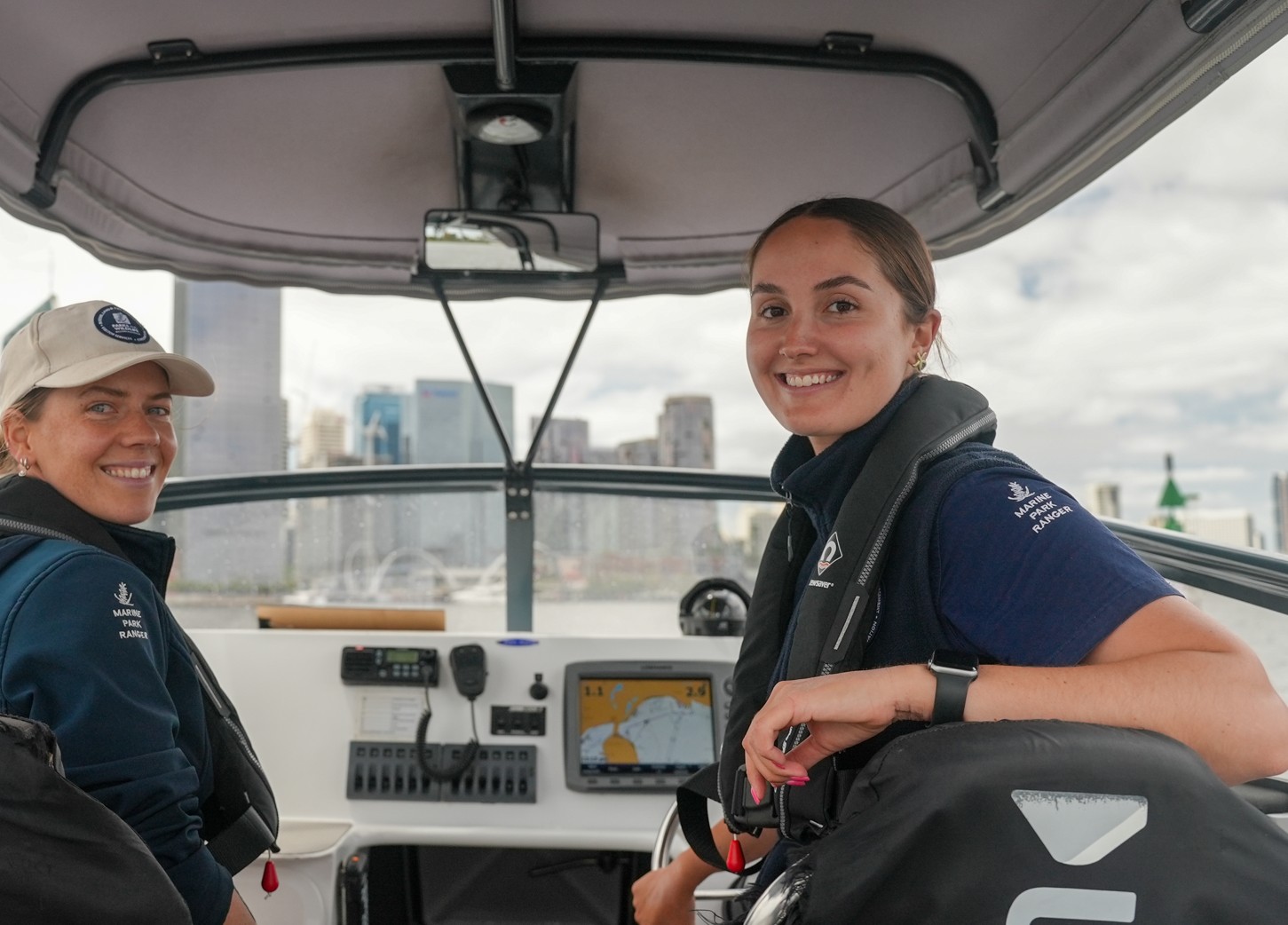 Two female Marine Rangers from WA's Department of Biodiversity, Conservation and Attractions shown at work on a patrol boat on the Swan River in front of the Perth city skyline.