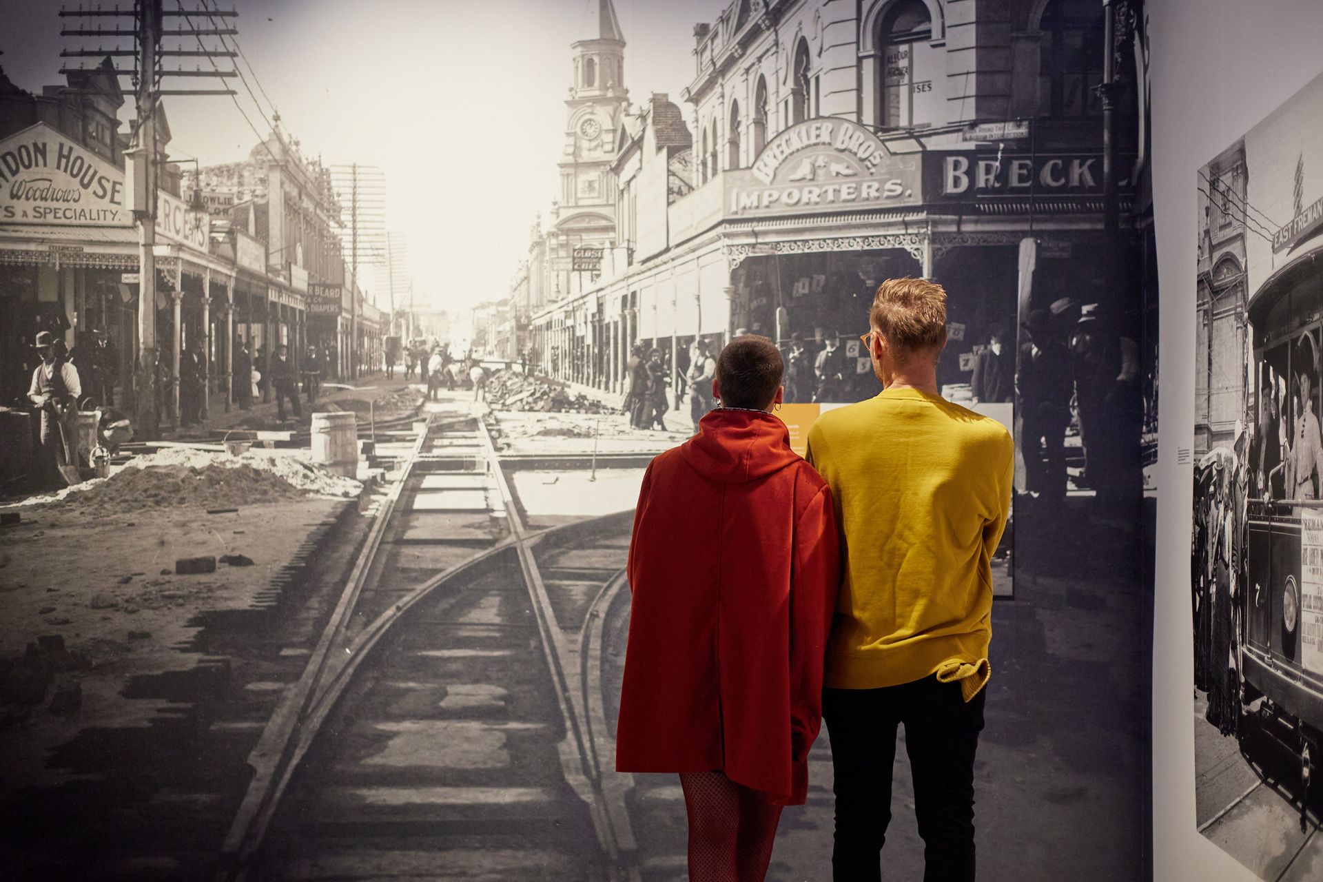 The image depicts two people with their backs to the camera staring at a large black and white historic photograph of workers building the Fremantle railway surrounded by old buildings.