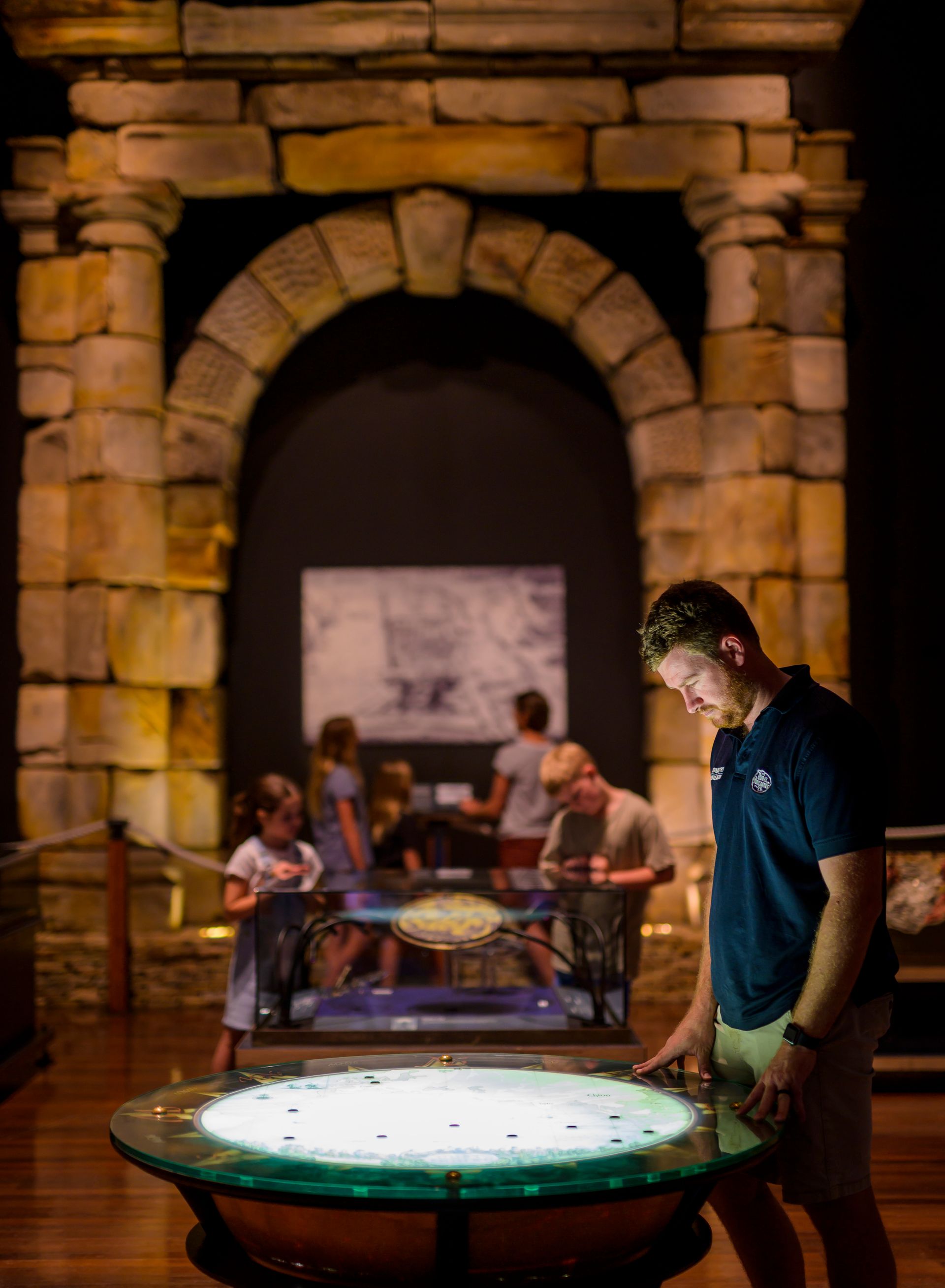 Image of a man in a navy t-shirt and green cargo shorts looking into a brightly lite screen with a limestone arch in the background.