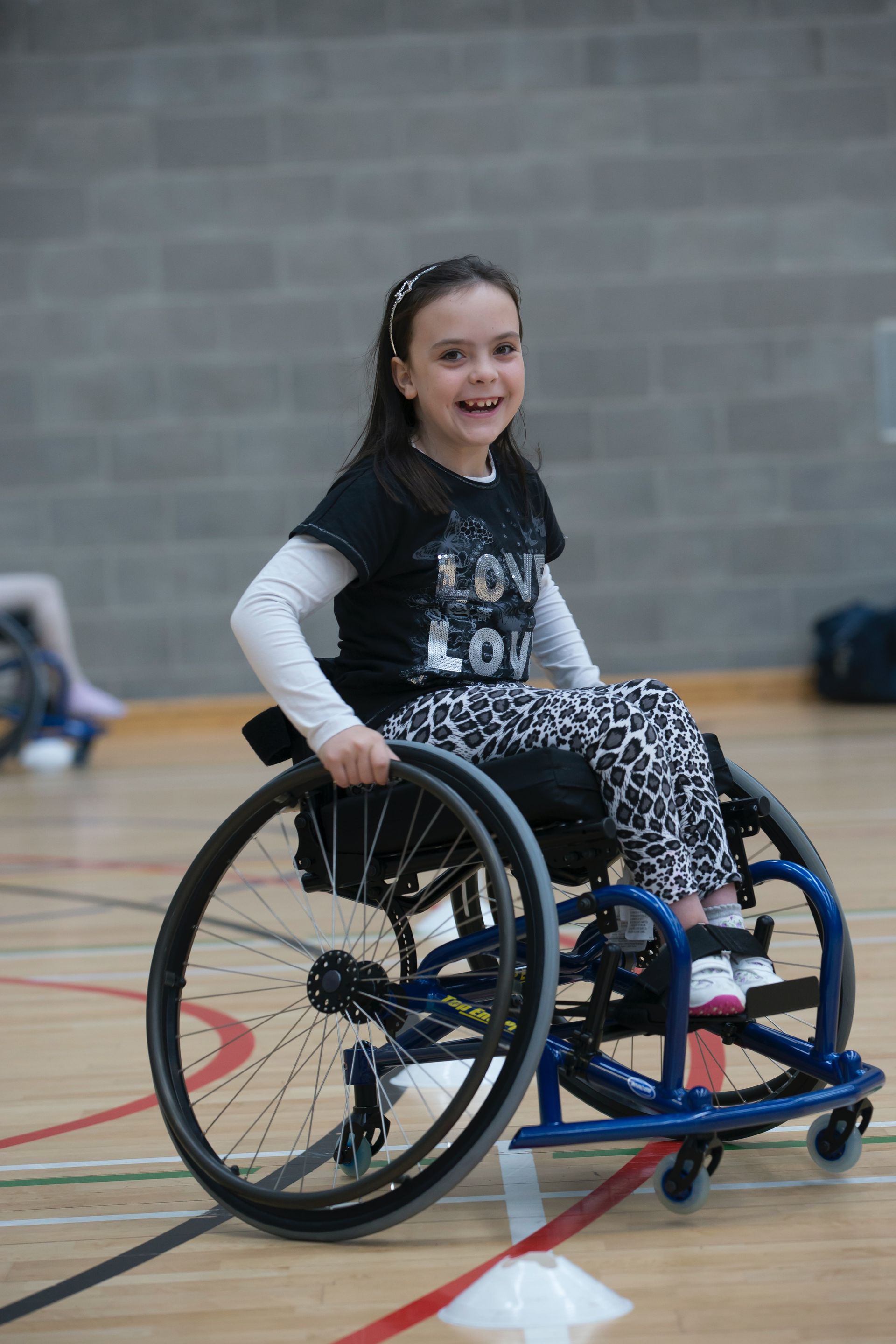 Girl in wheelchair and holding the wheels and smiling. Grey brick wall pictured in the background and a wooden floor with painted lines, indicating she is on a basketball court.