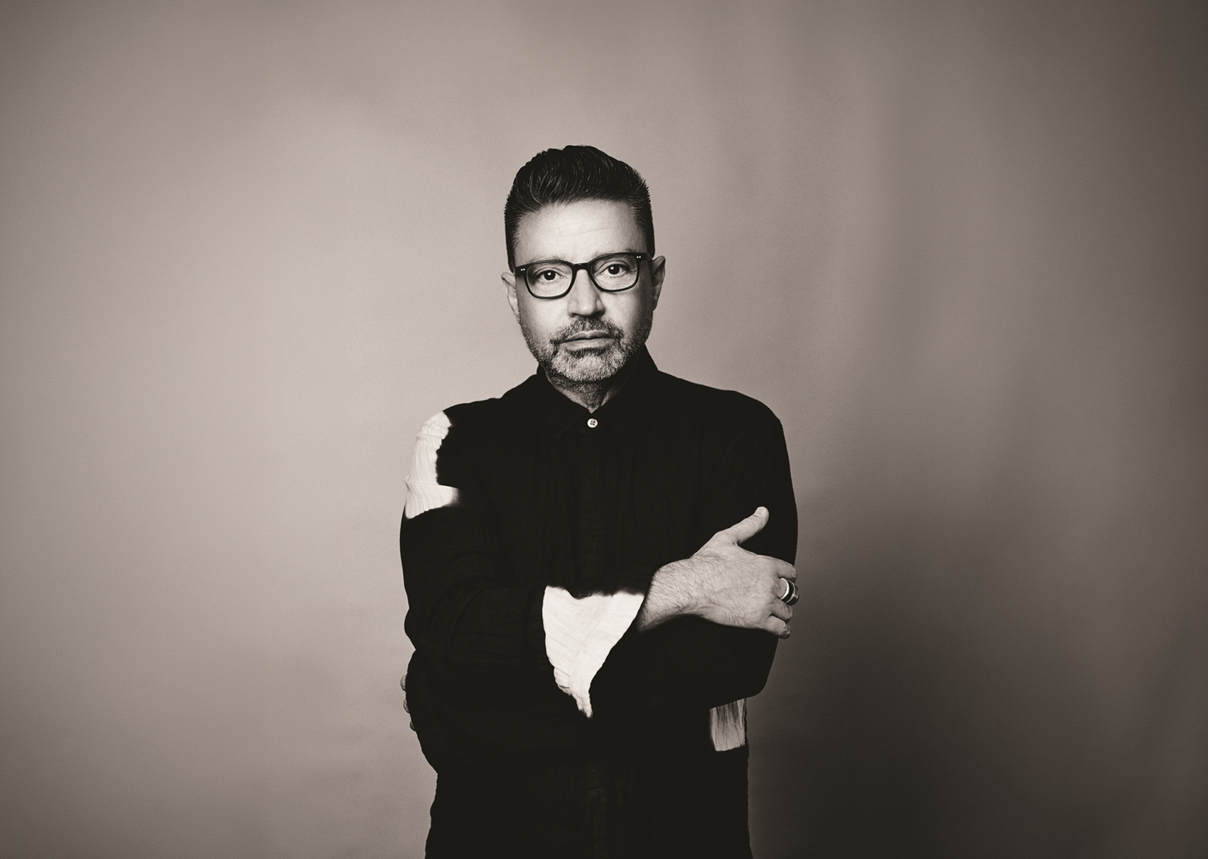 an adult person looks directly at the camera with a neutral expression. They stand with their arms almost crossed, wrapping their hands around the other arm. They are wearing dark framed glassed and dark coloured clothing with some light patterns.