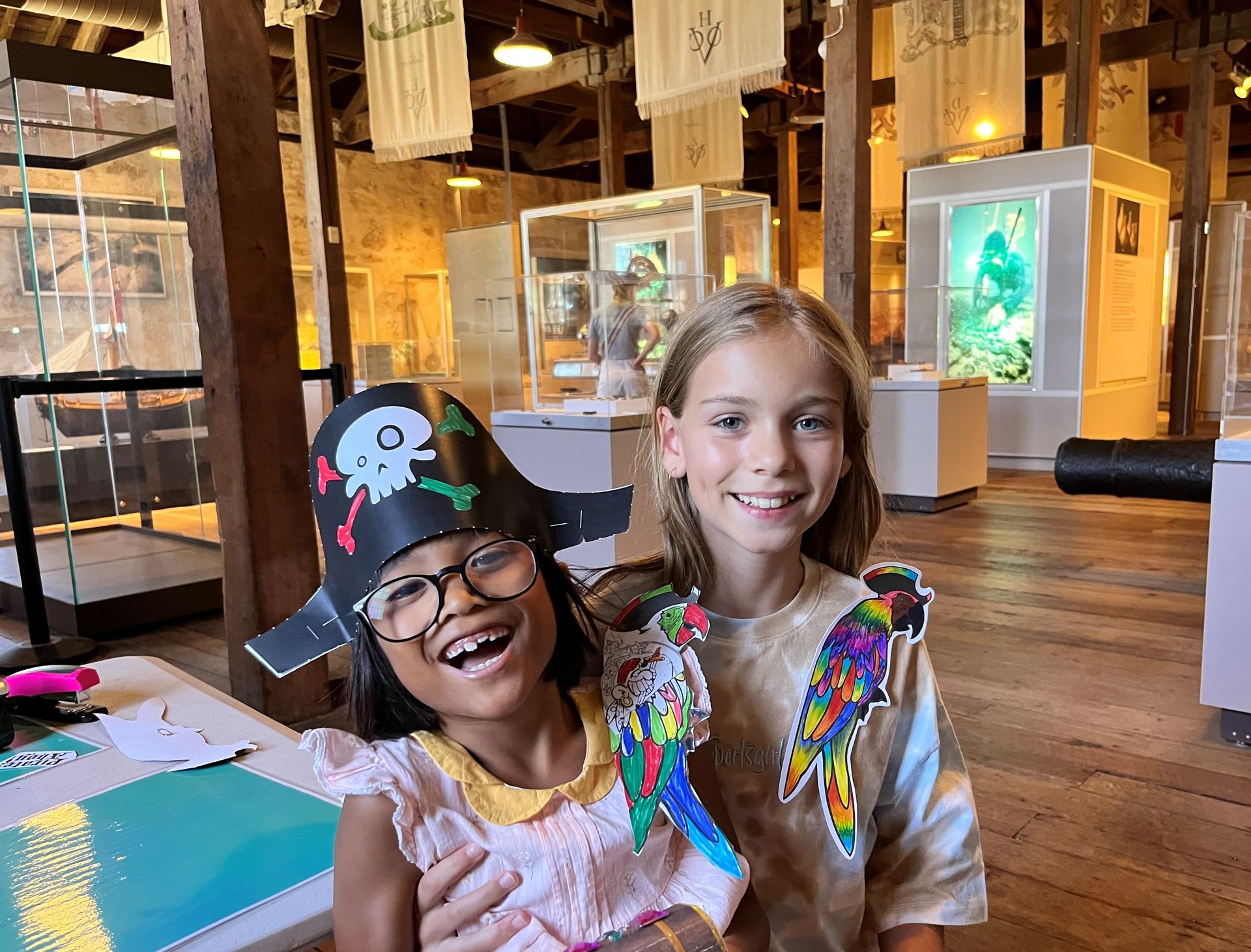 Two girls stand together smiling, wearing paper parrots on their shoulders. One is wearing a pirate hat too.  Behind them are display cases, a lightbox with a diver and historical flags are hung from the roof. The floor is wooden and the walls made of limestone.