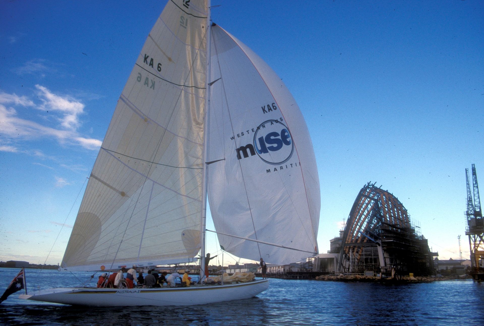The Australia II yacht, with a long off-white sail, and a WA Museum branded sail, moves towards the Maritime Museum in Fremantle which appears to be underconstruction, dating the image to the late 1990s or early 2000s