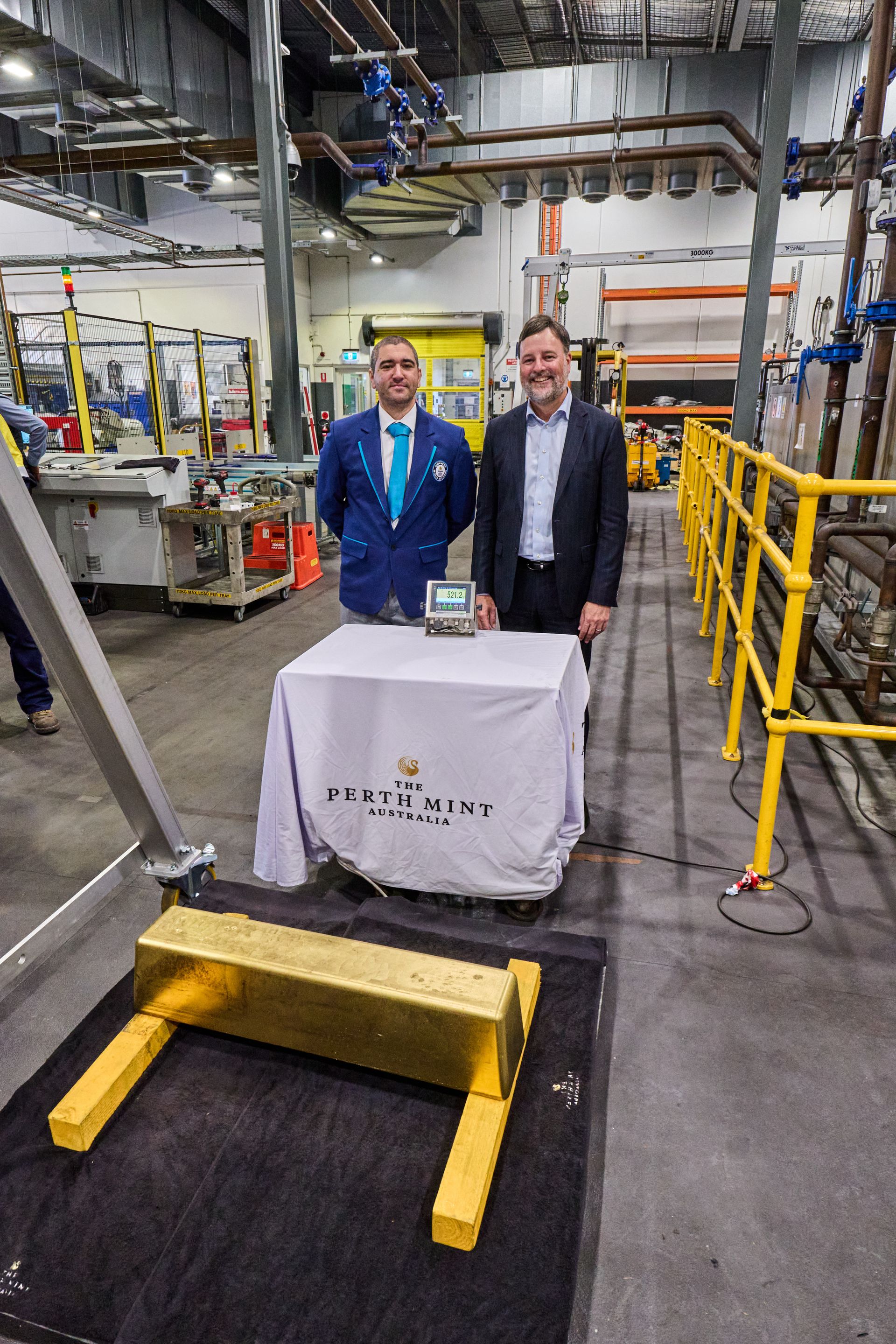 Two men in and industrial factory stand behind a large weighing scale where an enormous gold bar is positioned