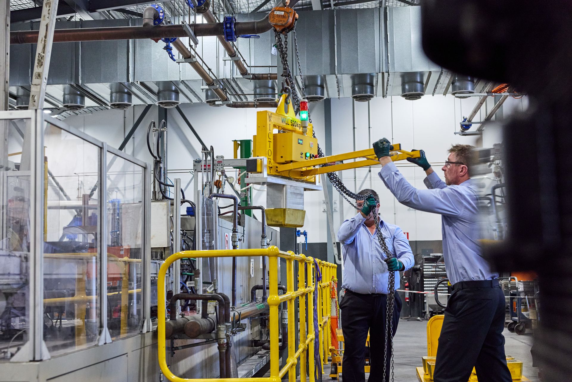 Two workers in an industrial factory use a vacuum lifter to lower a large gold bar into a mould