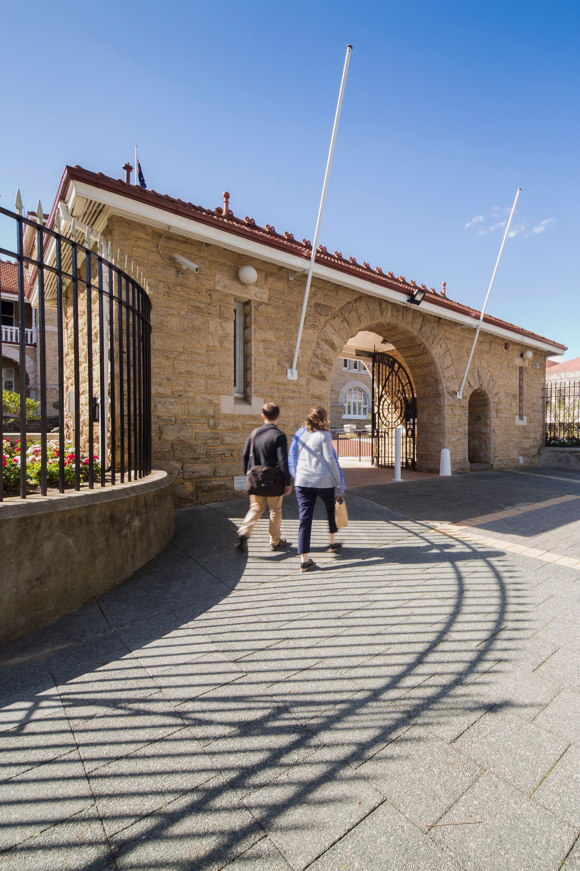 Symmetrical limestone gatehouse with orange tiles and arched entrance. The backs of two pedestrians are walking across pavement towards the gatehouse.