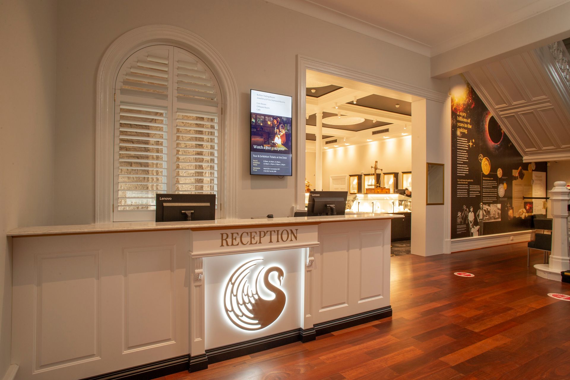 Interior of a building with white walls and timber flooring with louvered arched window. A long white reception desk in in the foreground that has a golden swan, the logo of The Perth Mint, positioned centrally.