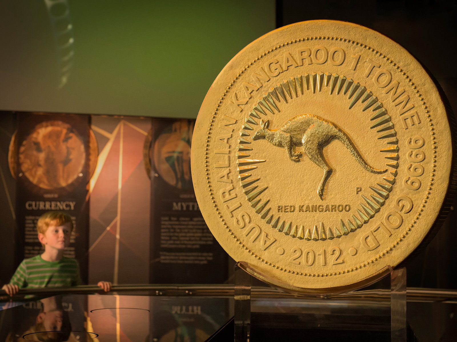 A young boy in a museum gallery looking at a large gold coin weighting one tonne. The coin has a jumping red kangaroo design in the centre with a circle of rays around it and the inscription 'Australian Kangaroo 1 Tonne 9999 Golf 2021'.