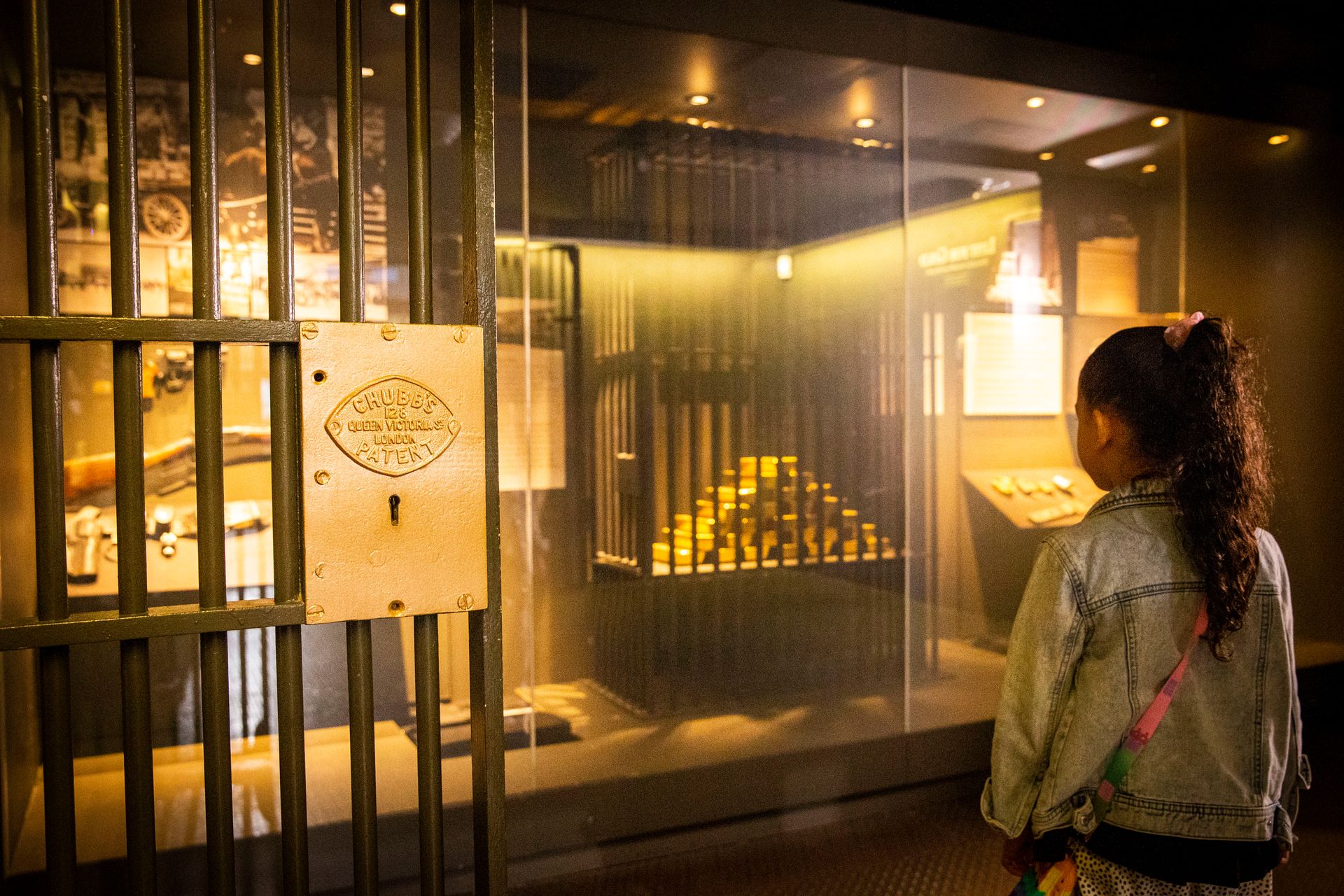 A child standing in a large walk-in vault looks at a display case containing a display of cast 12.44 kilogram gold bars inside a black barred enclosure.