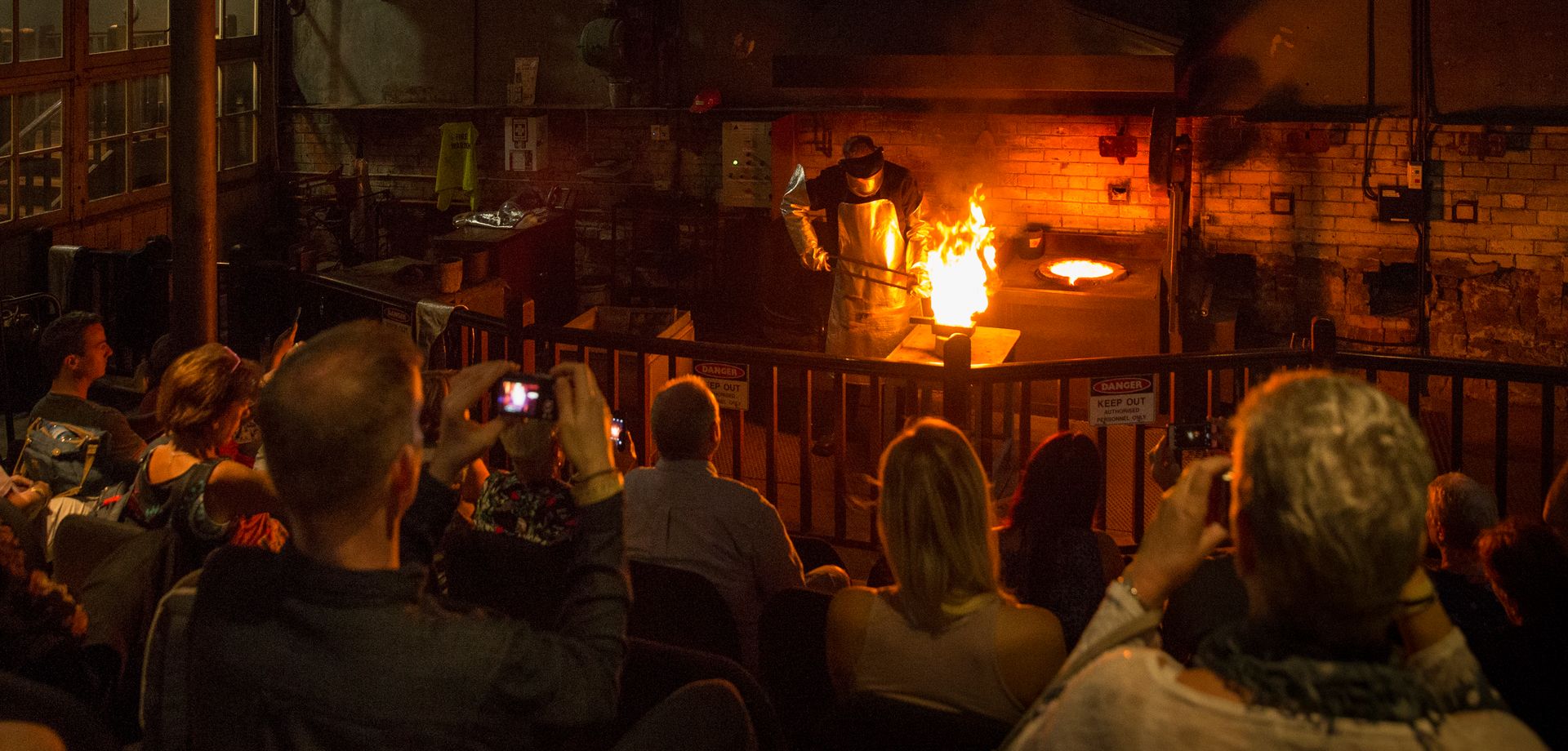 A group of people seated in a dimly lit room watch a worker wearing a blue uniform and personal protective equipment including a visor and a silver kevlar apron and gloves that extend up past the elbow. The worker is holds a hot ceramic crucible containing molten gold, that is glowing orange, with a long pair of pouring tongs. The cricule is tiled and flaming orange molten gold is pouring into a rectangular metal mould positioned on the bench. A lit furnace is in the background.