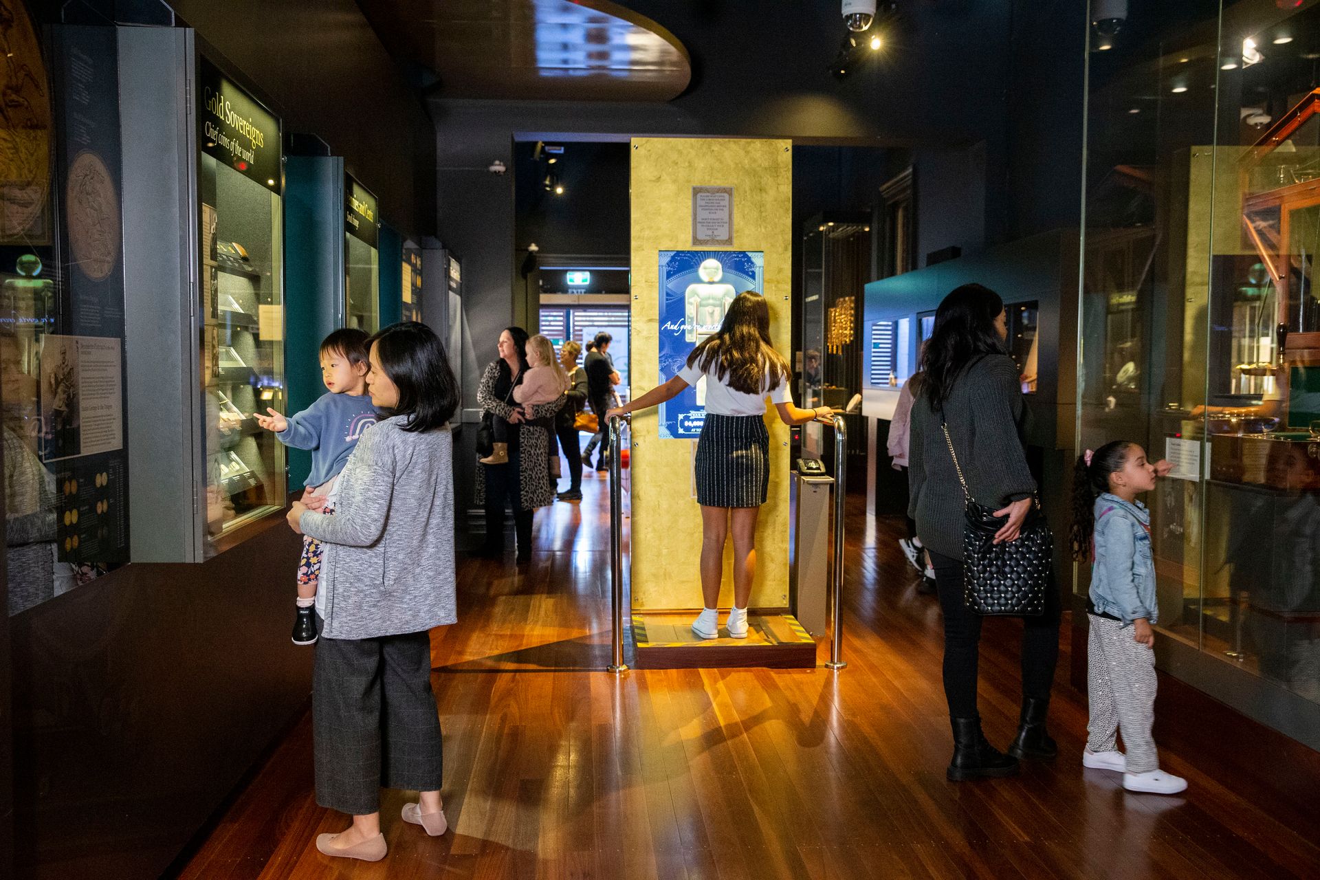 People looking at displays in a museum gallery with display cases on each side of the room and an child standing on an interactive scale that can tell her how much she is worth in today's gold price an