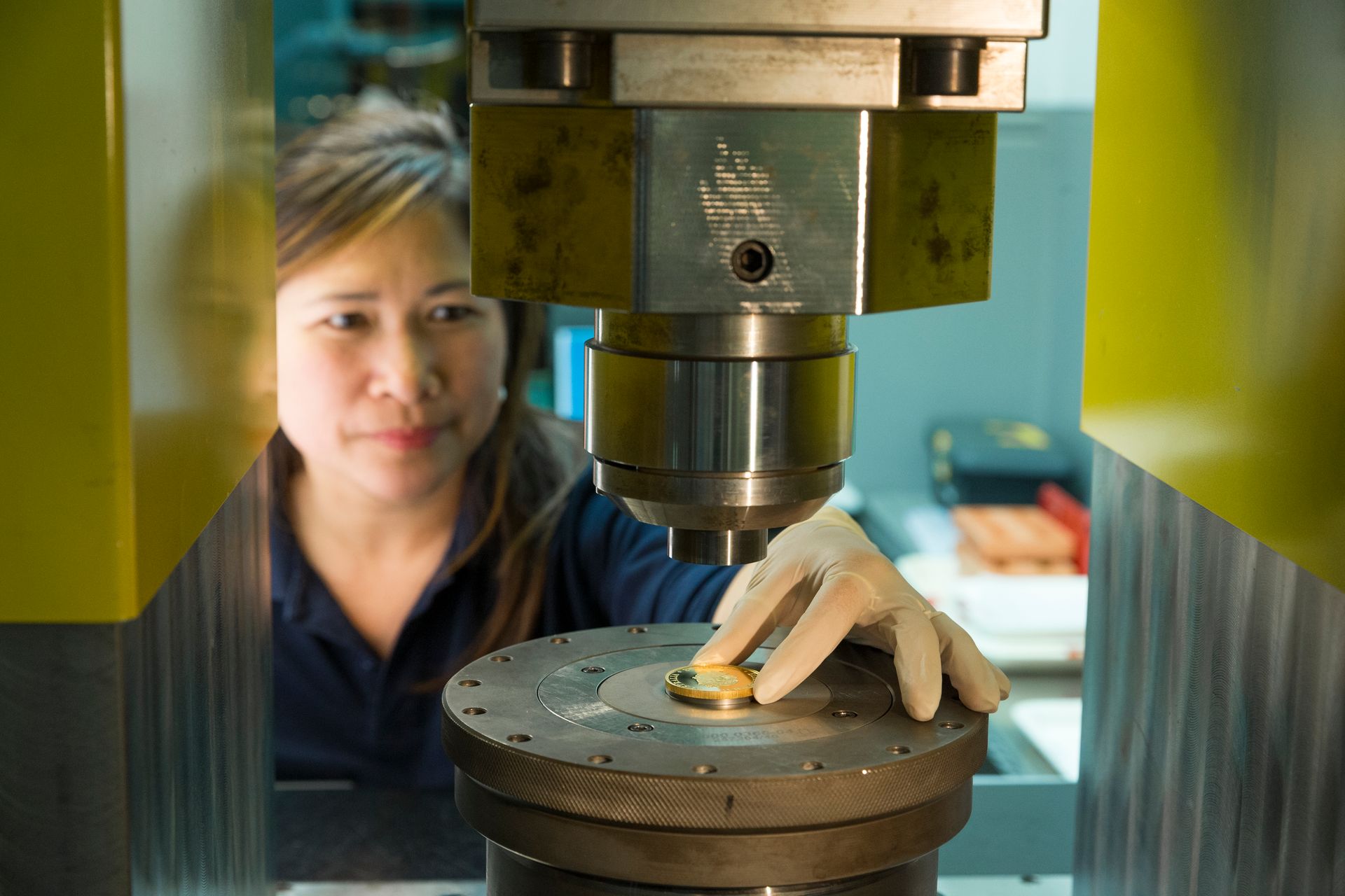 A female worked dressed in a blue uniform and wearing gloves is about to pick up a gold coin from the centre of a coin press that has been struck with a kangaroo design/